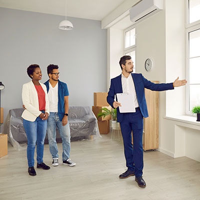 Un agent immobilier en costume montre et explique les caractéristiques d'un appartement vide à un couple, avec des cartons d'emménagement visibles en arrière-plan. 