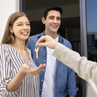 Couple souriant recevant un trousseau de clés des mains d'une tierce personne, symbolisant la remise des clés d'une nouvelle maison. 